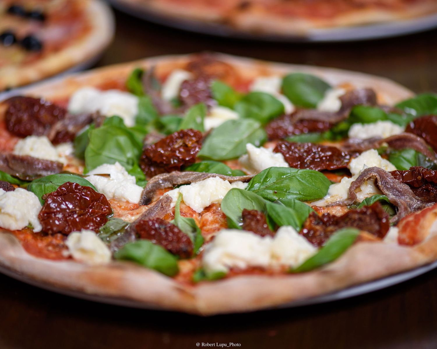 Freshly baked pizza topped with sun-dried tomatoes, fresh basil, white cheese, and anchovies on a dark wooden table.