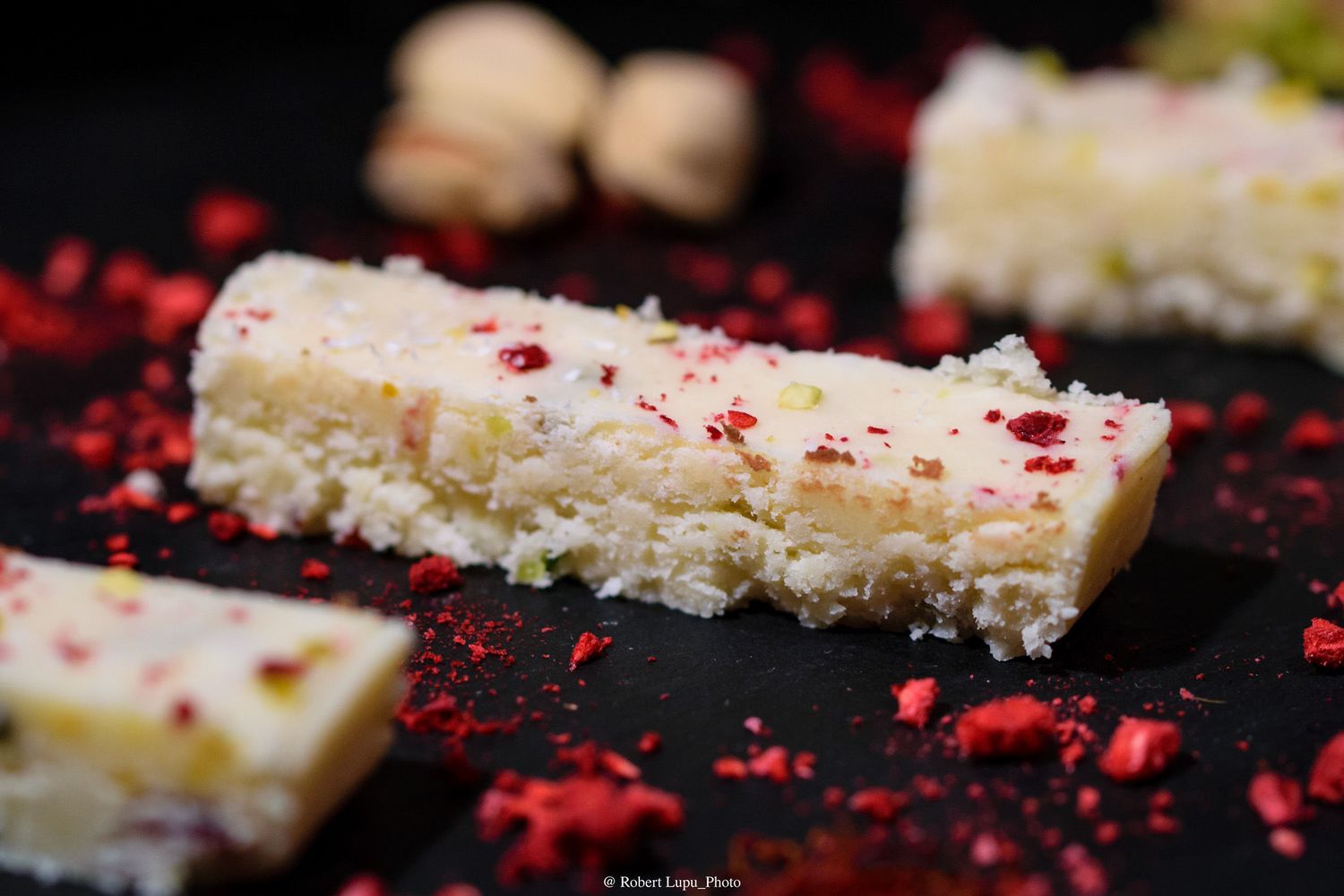 Close-up of white chocolate fudge bars topped with red raspberry crumbles and pistachios on a dark slate surface.