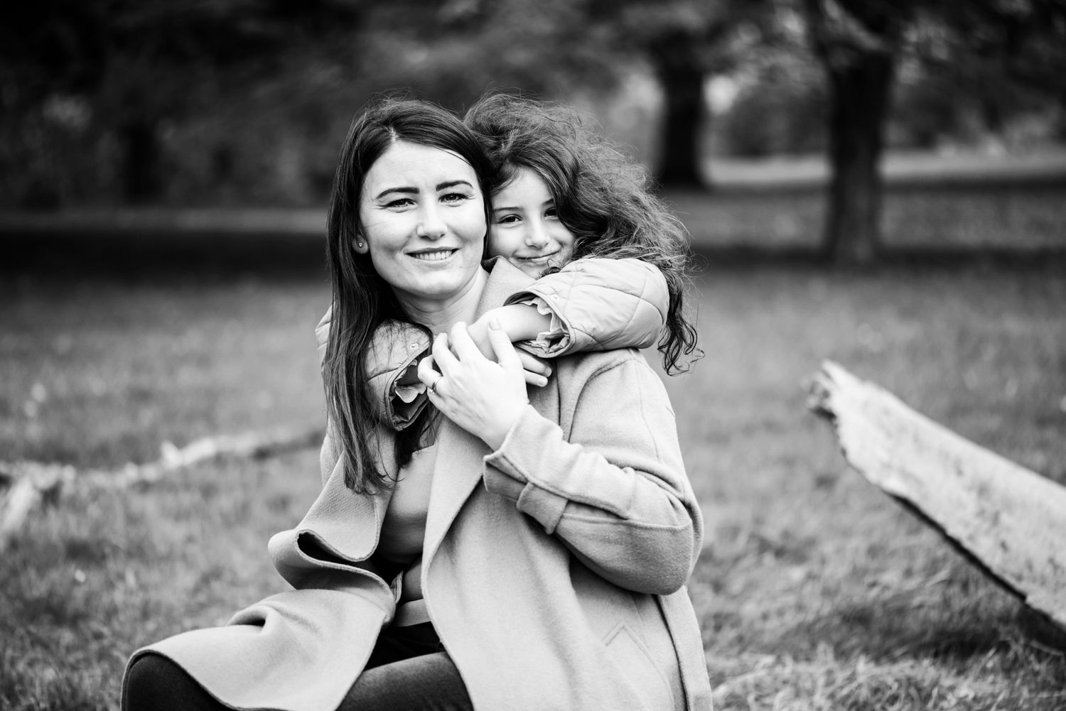 Black and white photo of a smiling woman and young girl hugging outdoors in a park setting.