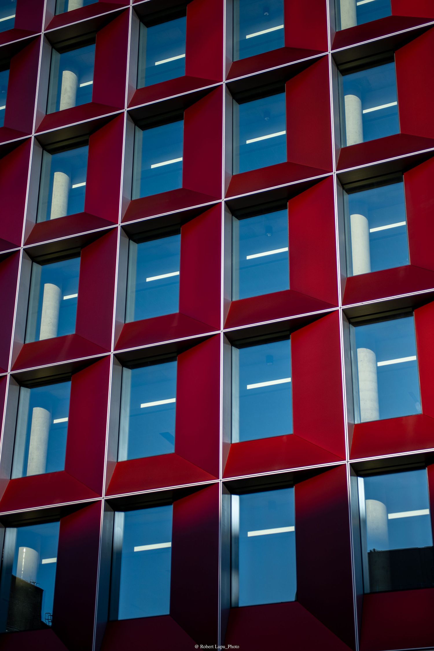 Red modern building facade with grid pattern of blue reflective windows showing white column reflections in natural light.