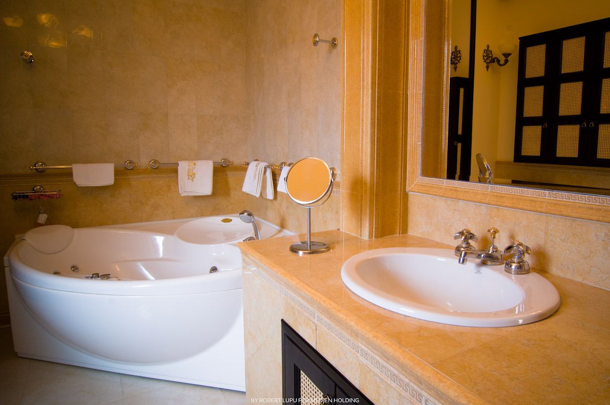 Luxury hotel bathroom featuring a white jacuzzi tub, marble vanity, round mirror, and warm beige stone walls.