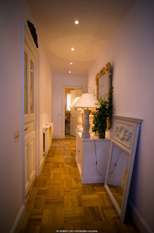 Elegant hallway with parquet flooring, white console table, ornate mirror, and glowing lamp leading to a bright room.