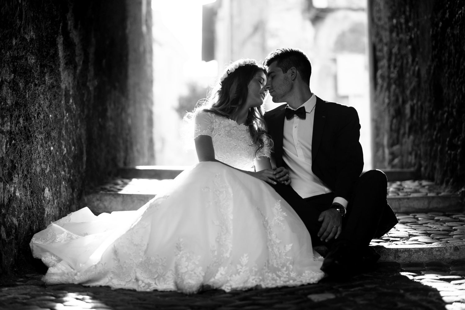 Black and white portrait of bride and groom sitting together in a rustic stone archway.