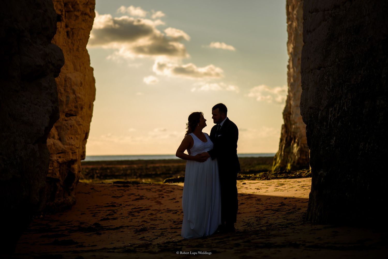 Bride and groom silhouetted between towering rock formations at golden sunset on sandy beach.
