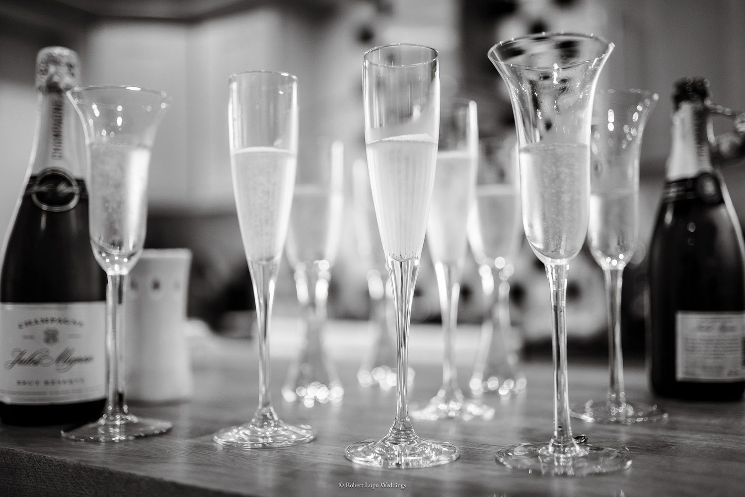 Black and white photo of elegant champagne flutes filled with sparkling wine beside bottles on a bar.