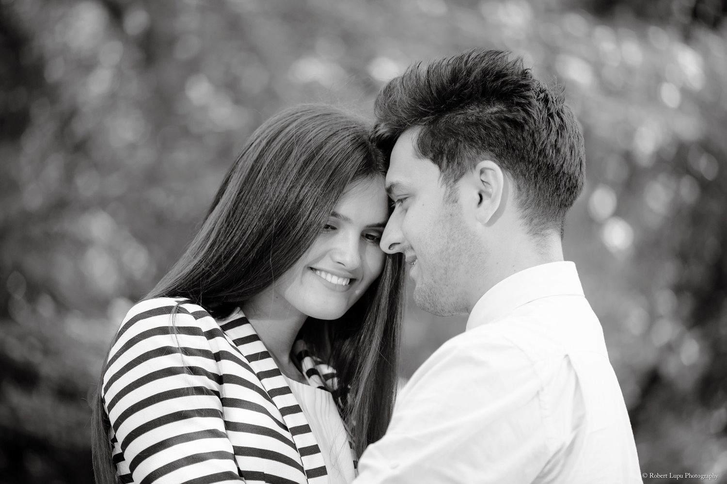 Black and white romantic photo of smiling couple touching foreheads outdoors among lush greenery.