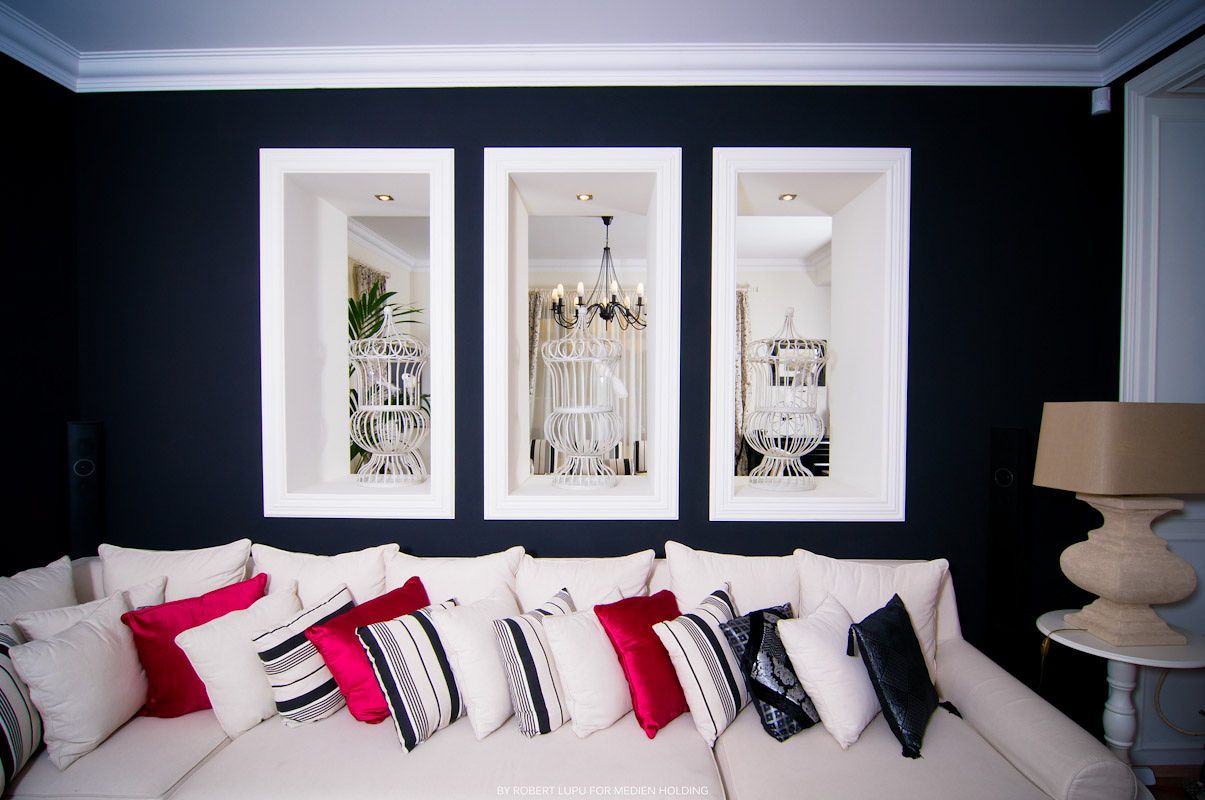 Elegant living room with navy blue walls, white sofa, colorful pillows, and three framed mirrors reflecting a chandelier.