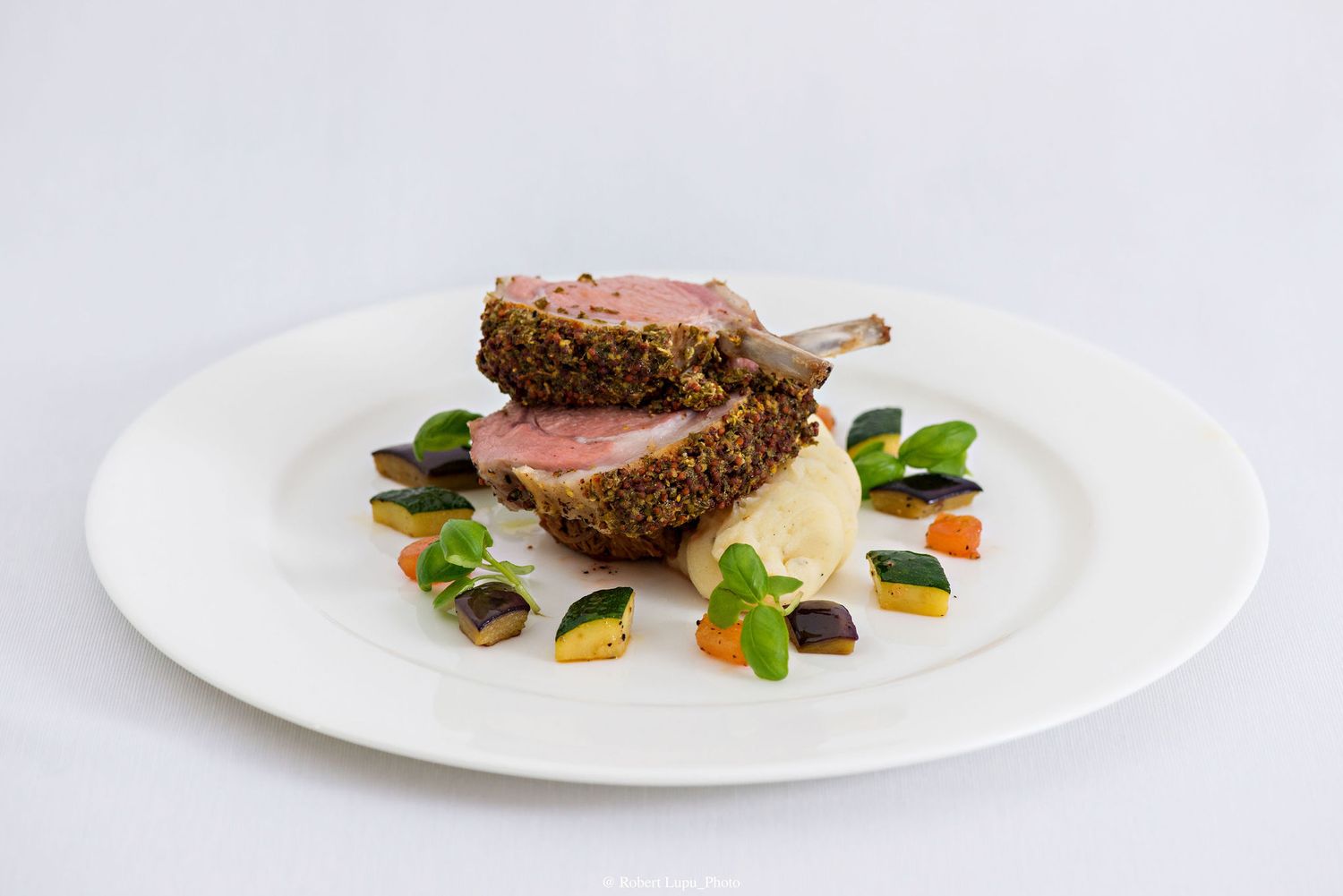 Herb-crusted rack of lamb with roasted vegetables and mashed potatoes on a white plate.