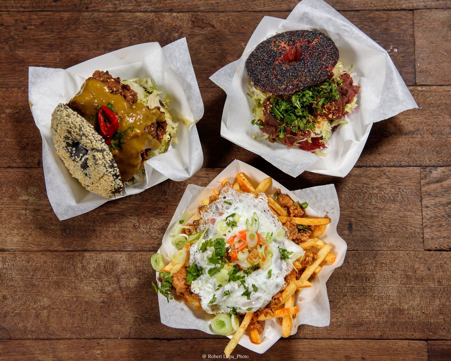 Three gourmet street food dishes on wooden table: loaded fries, black bun burger, and a sesame bun sandwich in paper trays.