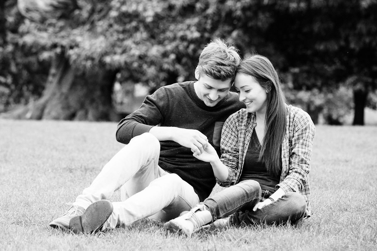 Young couple sitting on grass in park, sharing a tender moment together in black and white photo.