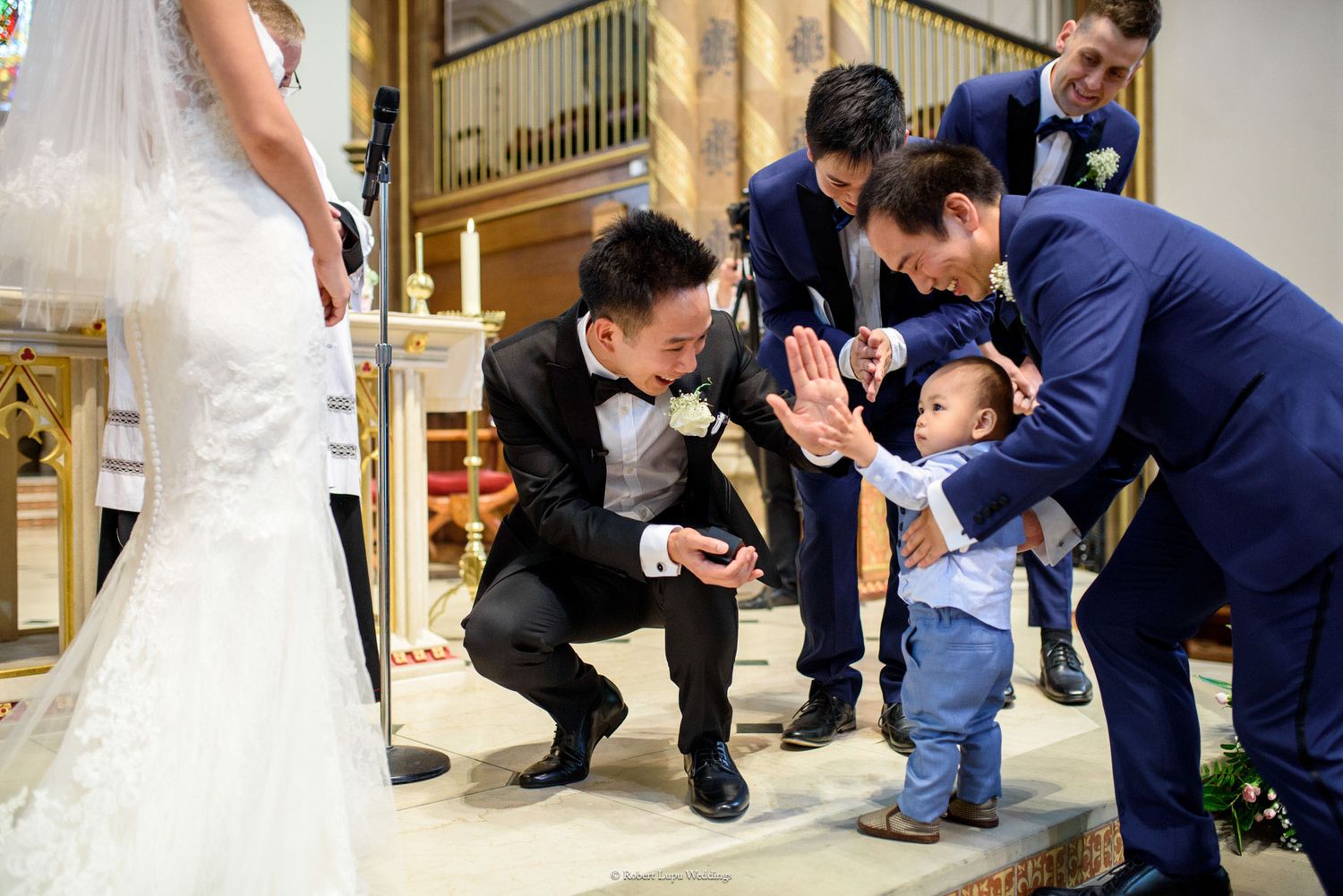 Groom crouches to high-five a toddler at a wedding ceremony inside a church, with groomsmen smiling nearby.