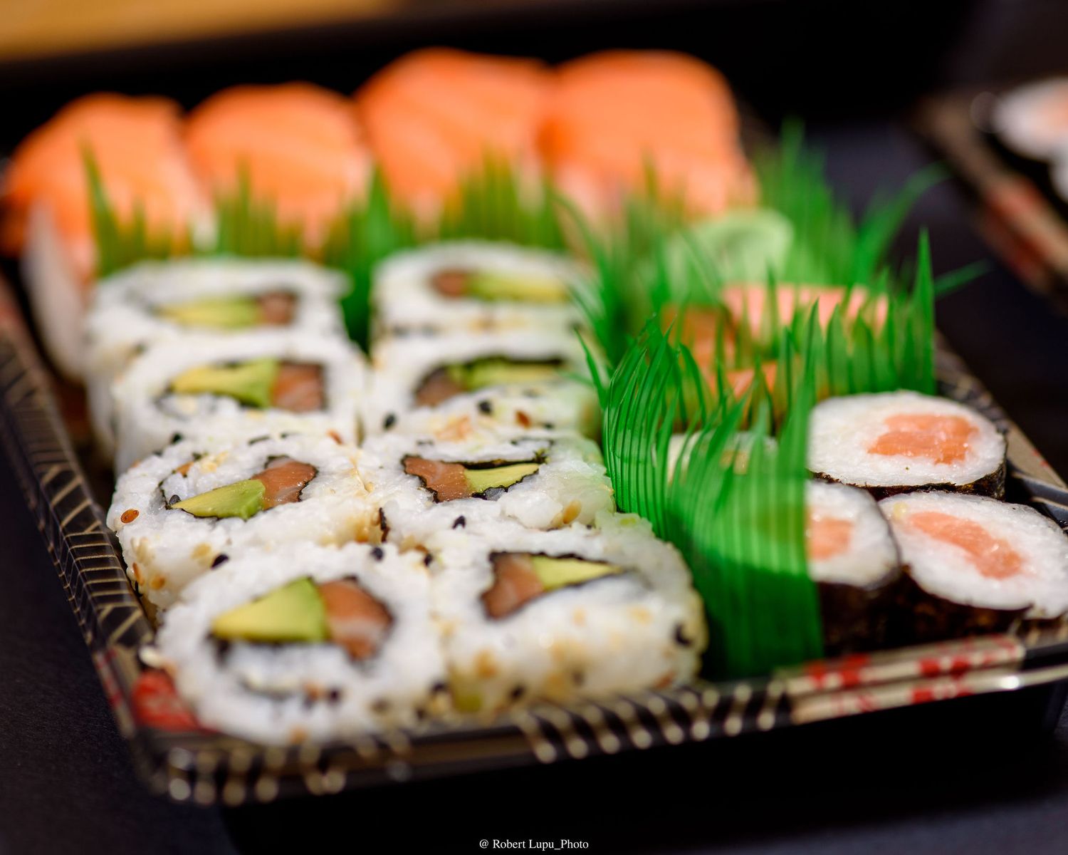 Assorted sushi platter with California rolls, salmon nigiri, and maki rolls on a black tray with green garnish.