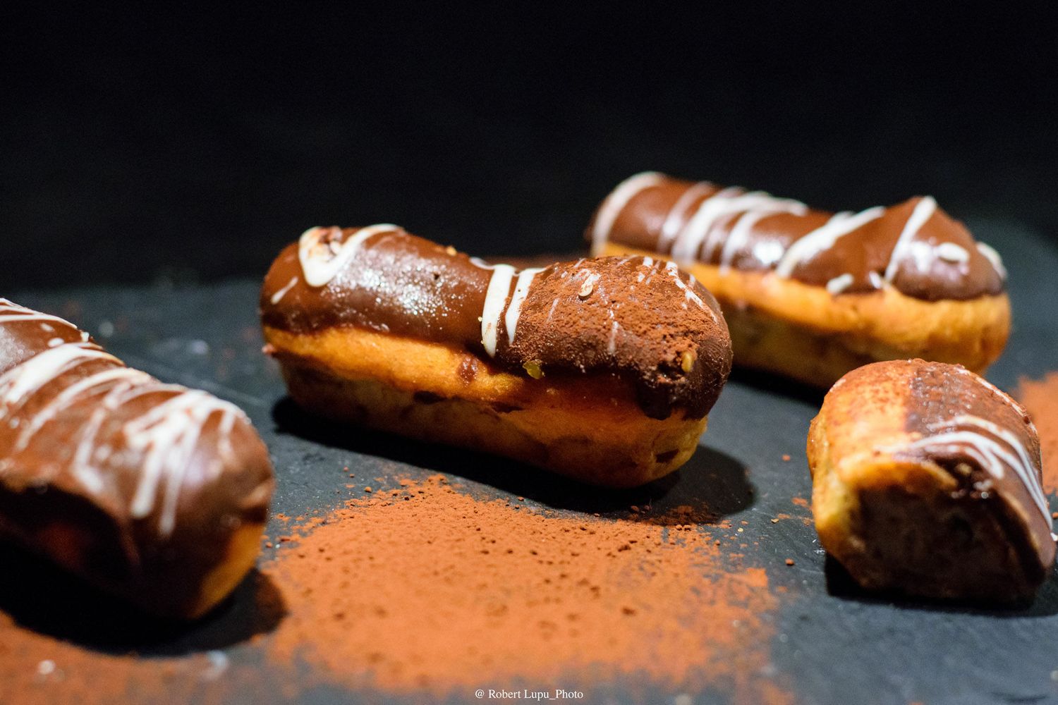 Chocolate eclairs dusted with cocoa powder and drizzled with white chocolate on a dark slate surface.
