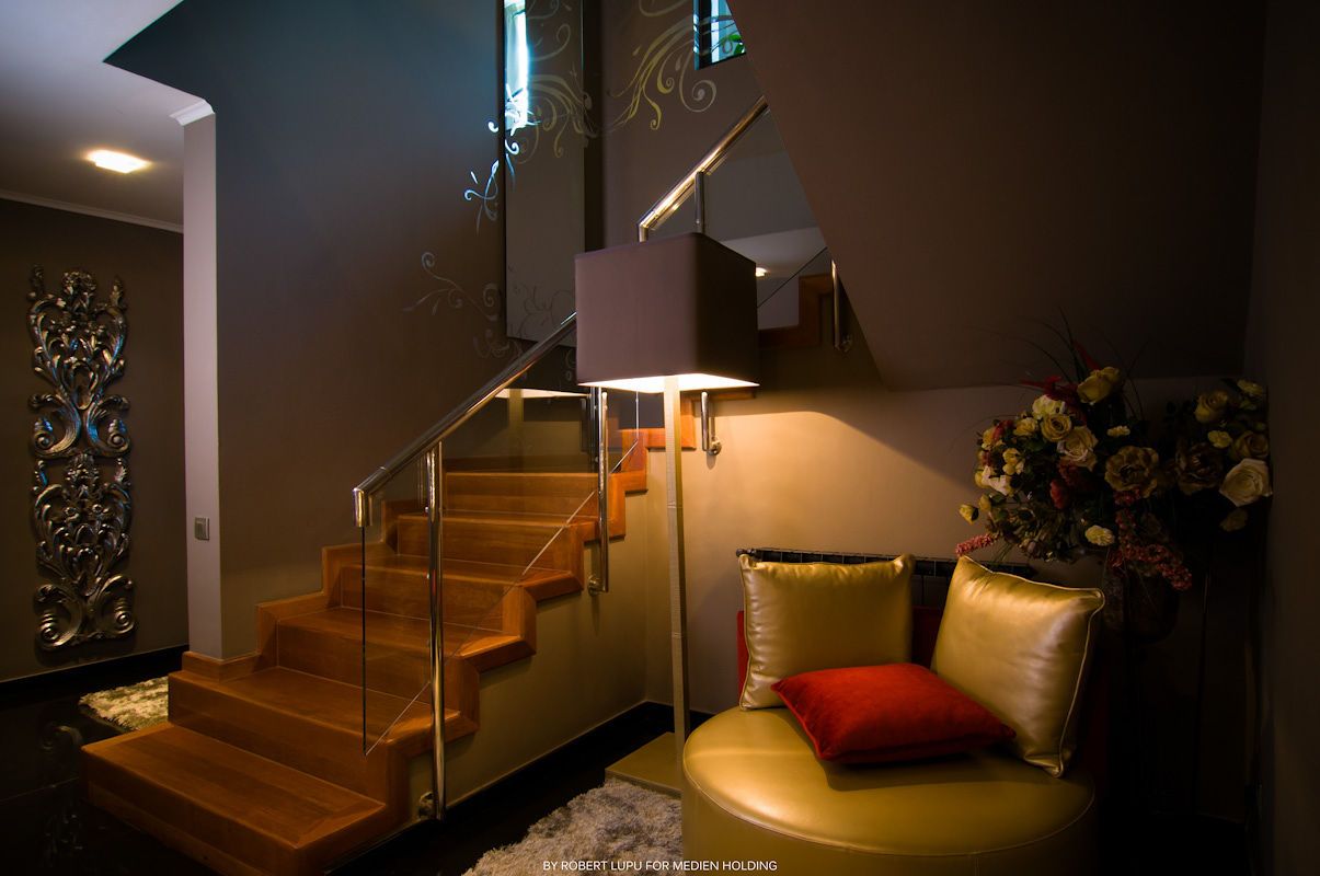 Modern interior with wooden staircase, glass railing, warm lighting, and cozy seating area with colorful pillows.
