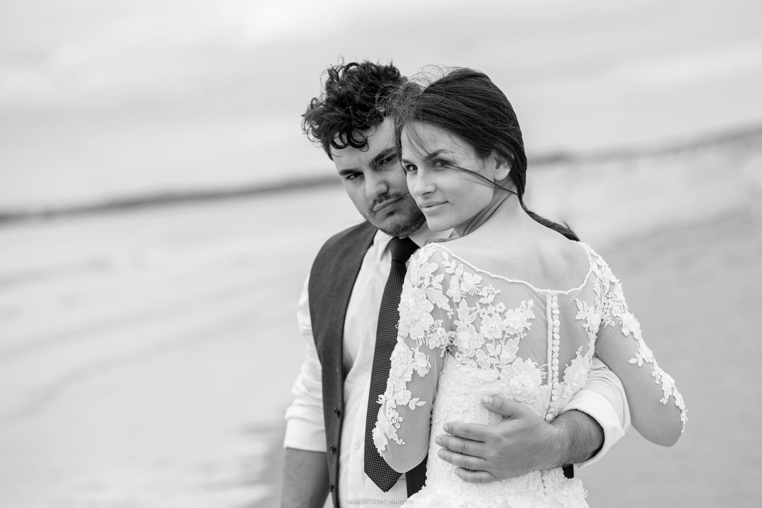 Bride in lace wedding dress and groom in dark suit embrace romantically on sandy beach in black and white photo.