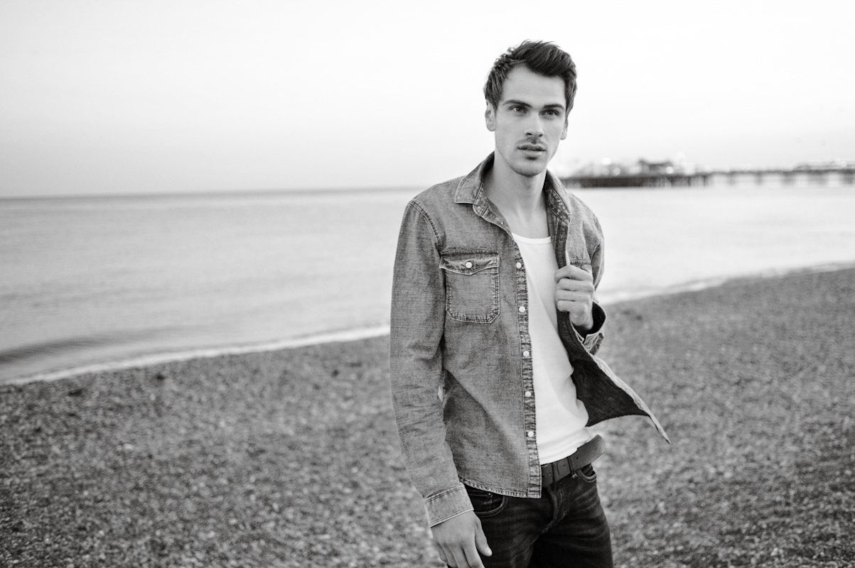 Young man in denim shirt standing on pebble beach with pier in background, black and white photography.