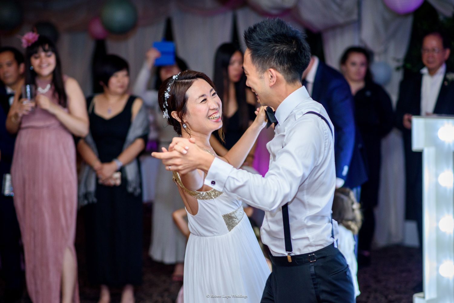 Bride and groom share first dance at wedding reception with guests watching in background.
