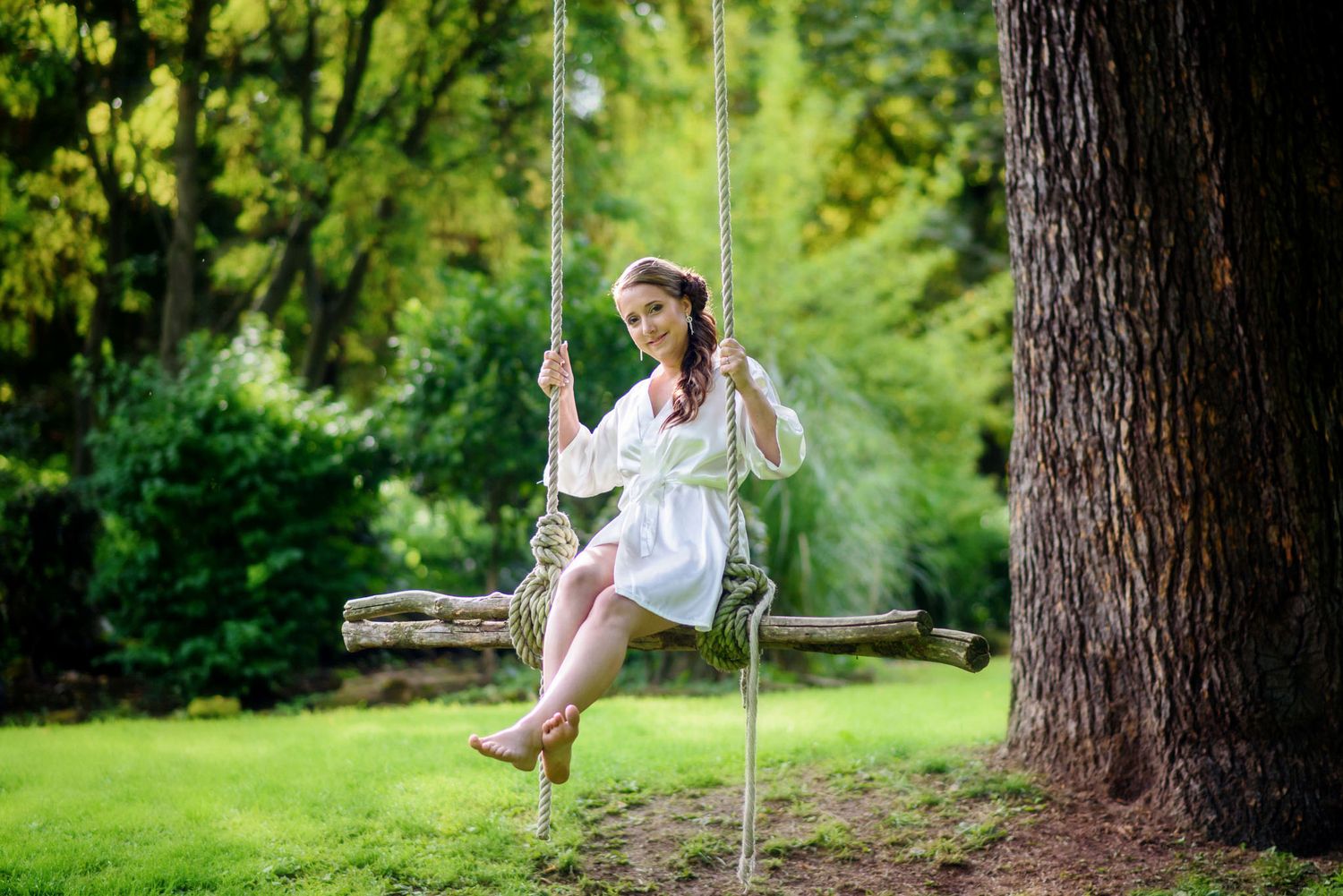 Young woman in white dress sitting on a rope swing attached to a large tree in a lush green garden.