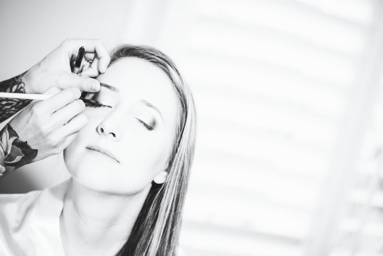 Black and white photo of a woman getting eye makeup applied by a makeup artist with a brush.