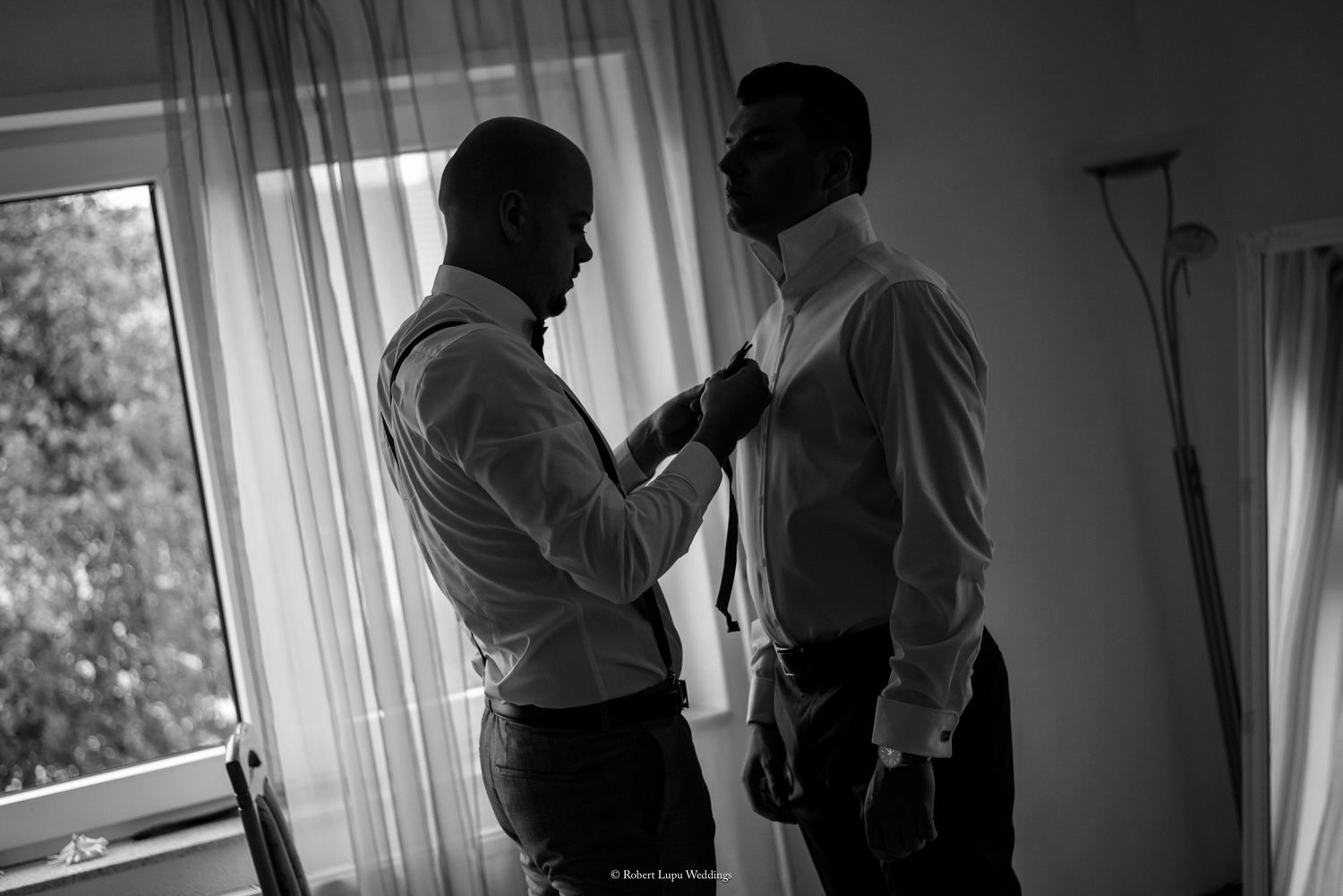 Black and white photo of two men in white dress shirts, one helping the other adjust a tie near a bright window.