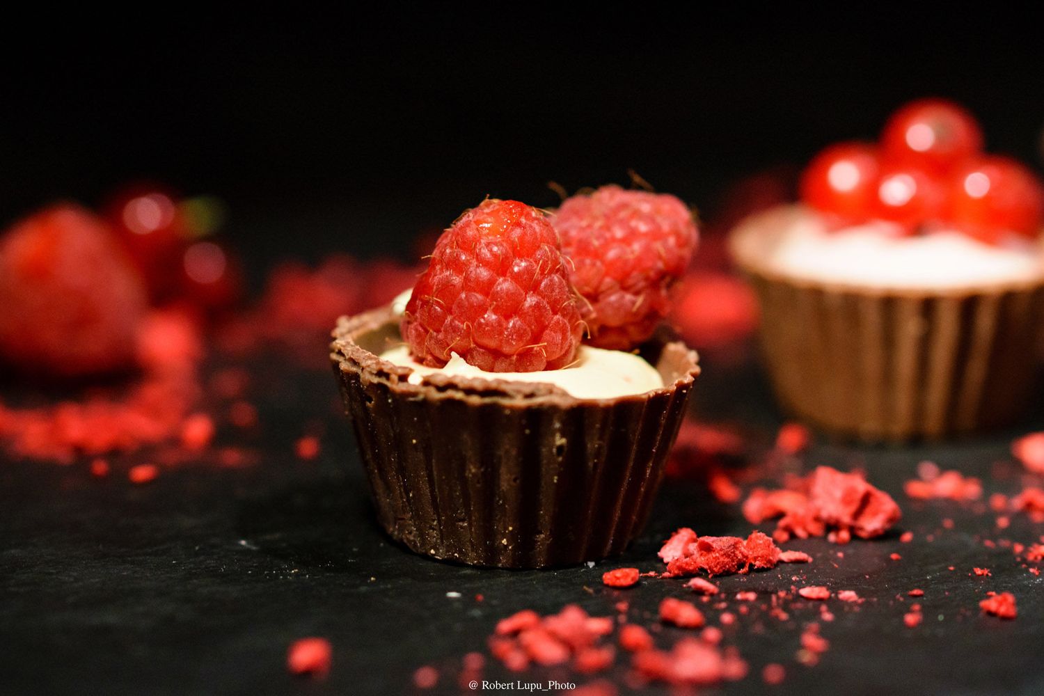 Chocolate cup dessert topped with fresh raspberries and cream, surrounded by red berry crumbs on a dark surface.