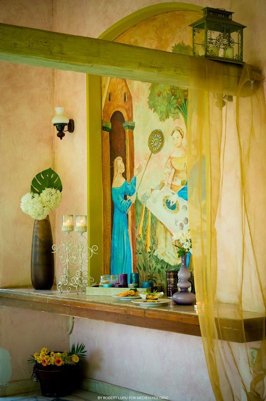 Elegant Mediterranean-style interior with a Renaissance mural, blue figurine, candles, and sheer yellow curtain on a decorative shelf.