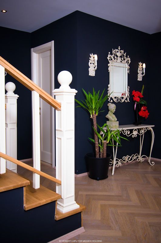 Elegant entryway with navy blue walls, white staircase railing, ornate mirror, bust sculpture, and tropical plants on herringbone floor.