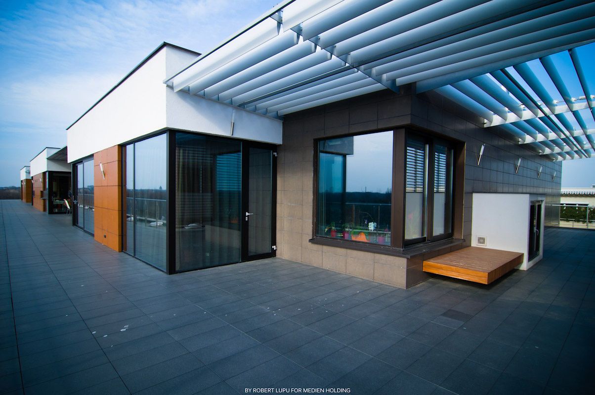 Modern rooftop terrace with glass walls, wooden accents, and a pergola under a clear blue sky.