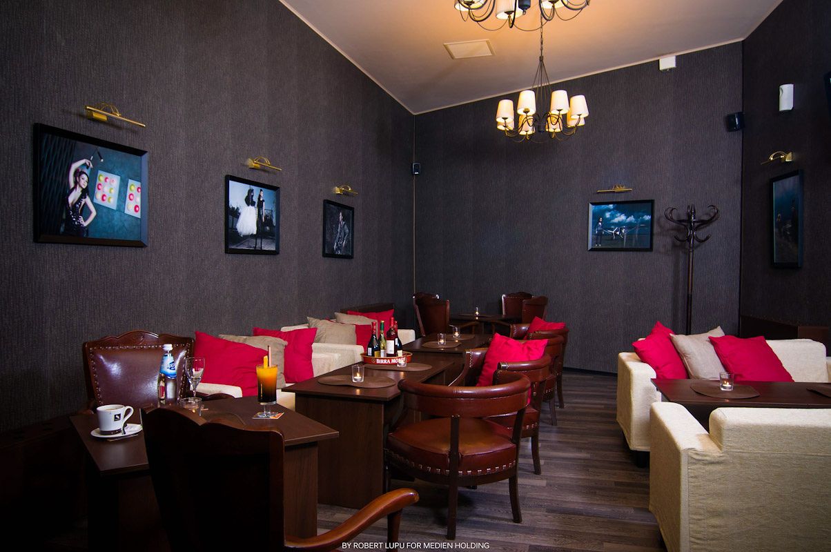 Cozy restaurant interior with dark walls, framed artwork, wooden tables, red accent chairs, and elegant chandelier lighting.