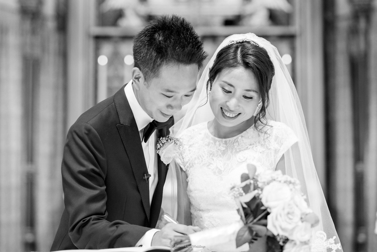 Black and white photo of a smiling Asian bride and groom signing their wedding certificate in an elegant venue.