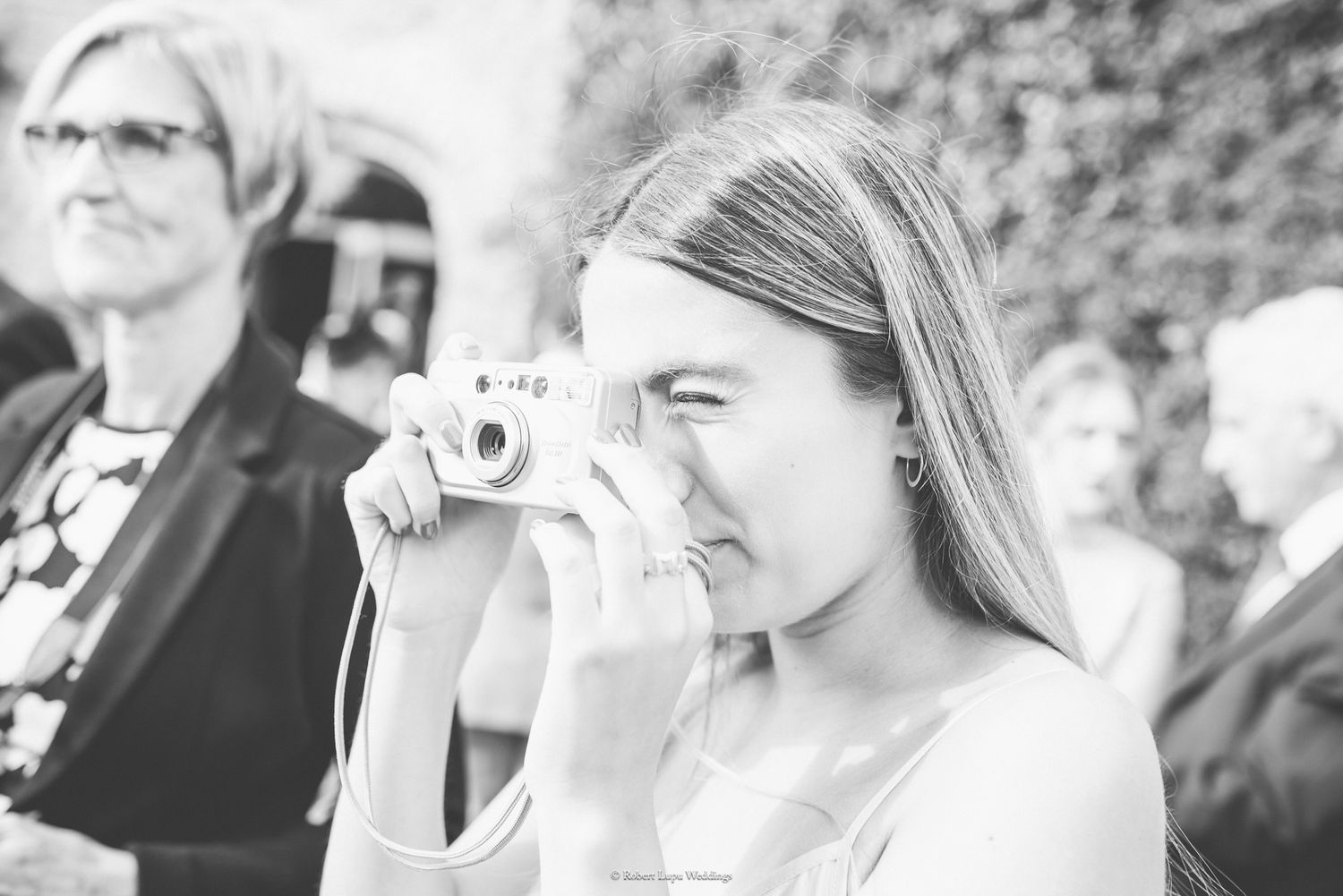 Young woman photographing with a vintage camera at an outdoor event, with guests visible in the background.