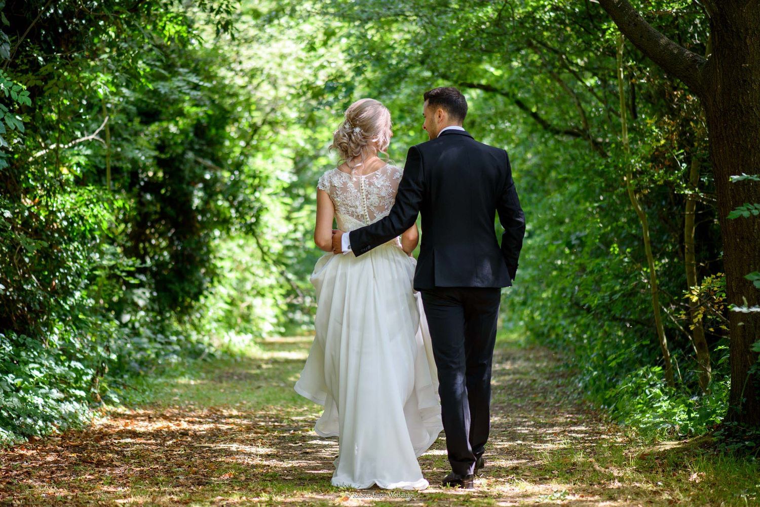 A bride and groom walk hand in hand down a sunlit tree-lined path on their wedding day.
