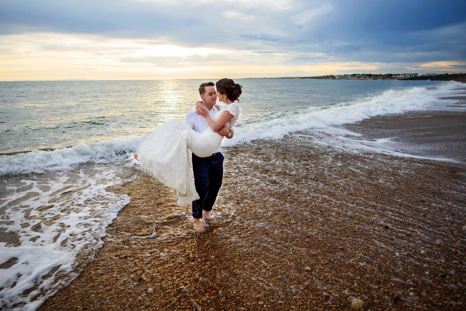 Groom carrying bride in white wedding dress along sandy beach at sunset with ocean waves.