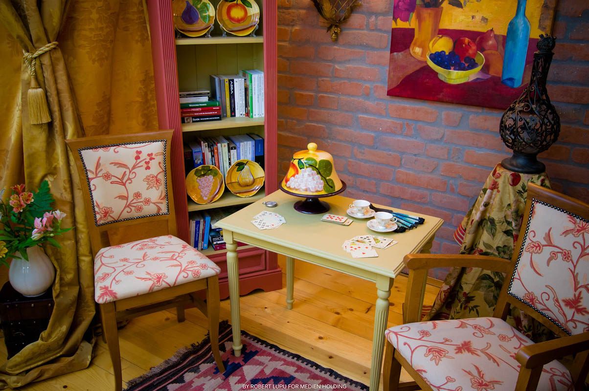 Cozy corner dining nook with floral chairs, wooden table, colorful bookshelf, and decorative artwork on brick wall.