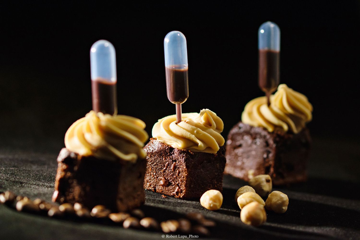 Chocolate brownie bites topped with yellow frosting and test tube syringes on a dark background.