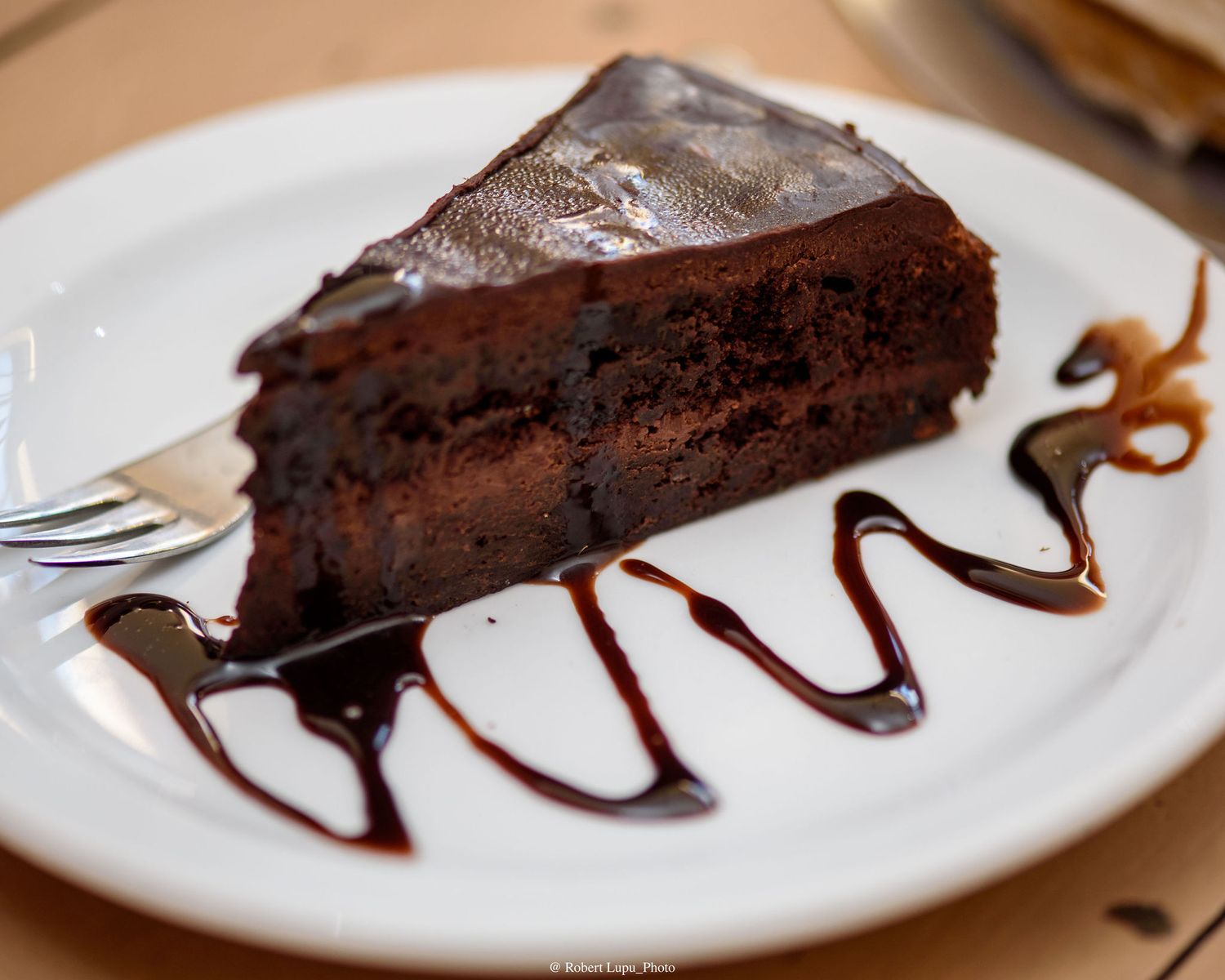 A slice of rich chocolate cake with glossy ganache topping and chocolate sauce drizzle on a white plate.