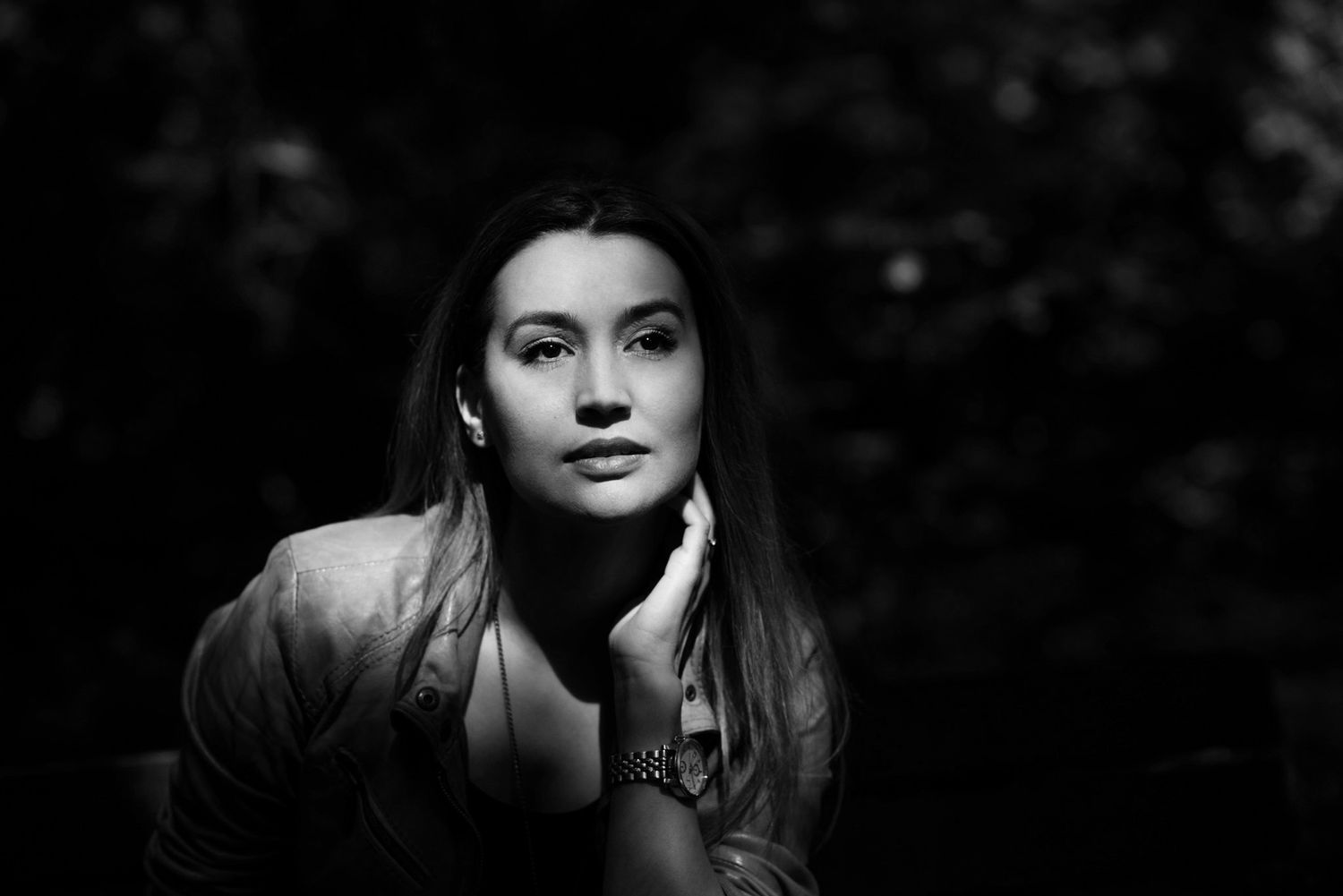 Black and white portrait of a young woman resting her hand on her chin, gazing thoughtfully in soft natural light.