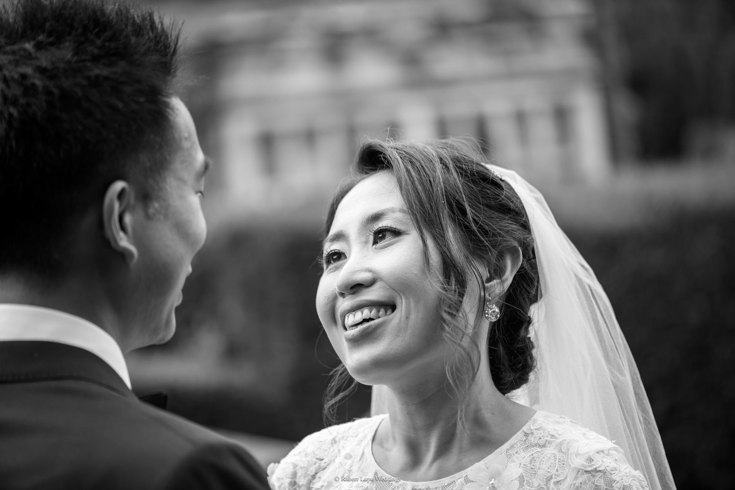 Black and white photo of a smiling Asian bride in veil gazing at groom during outdoor wedding ceremony.