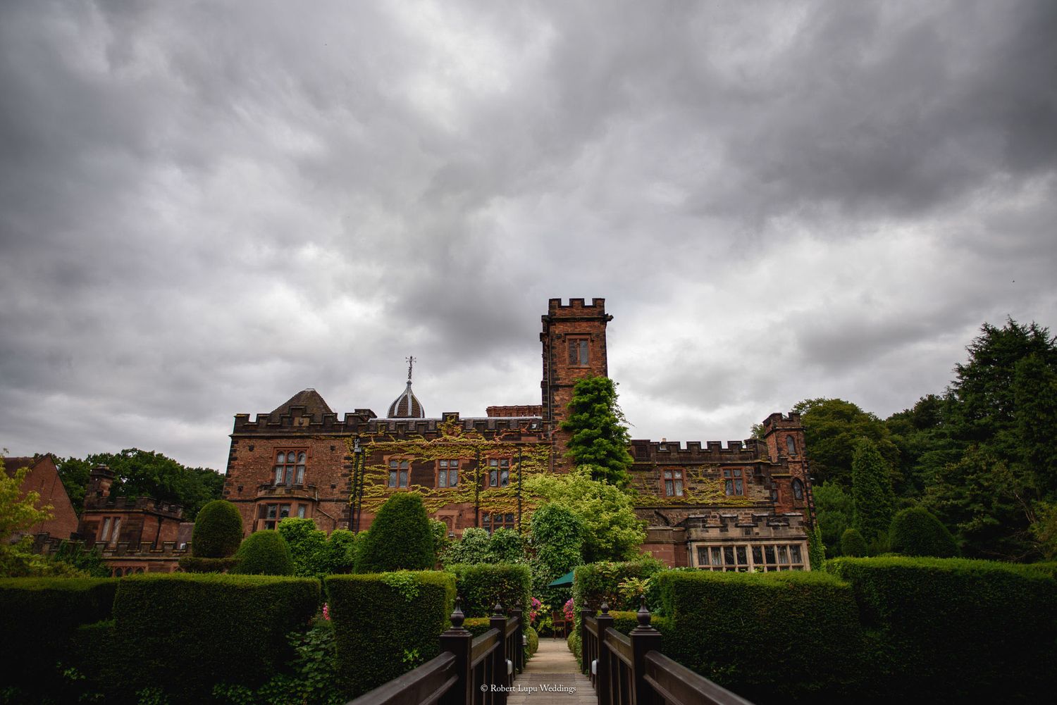 A grand red-brick castle with manicured topiary gardens under dramatic stormy skies in England.