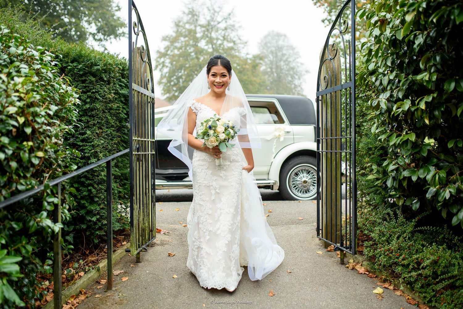 Bride in white lace gown and veil holding bouquet walks through iron gate with classic white car behind her.