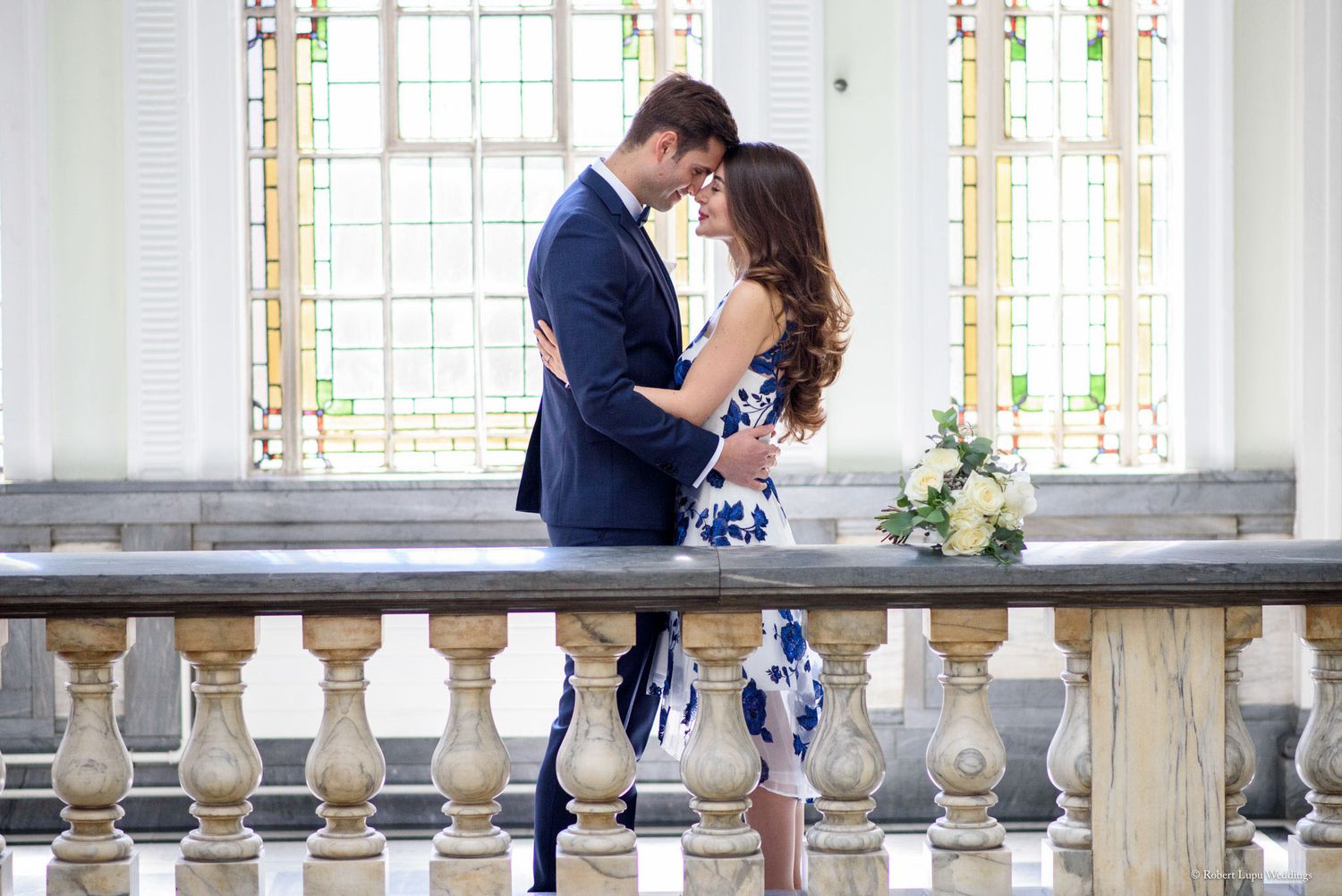 Couple embracing romantically on an elegant balcony with stained glass windows, man in navy suit, woman in blue floral dress.