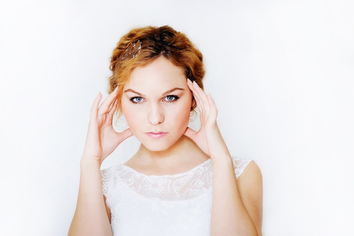 Young blonde woman in white dress holding temples with both hands, appearing stressed or having a headache.