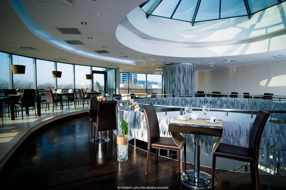Elegant rooftop restaurant with panoramic windows, round skylight, and formally set dining tables with brown chairs.
