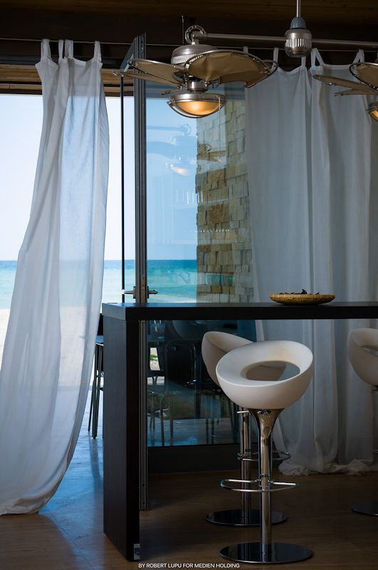 Modern bar stool beside a dark counter with white curtains and ocean view, featuring a ceiling fan in a coastal interior.