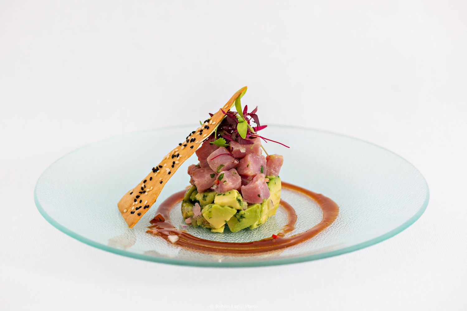 Elegant tuna tartare with avocado, microgreens, sesame cracker, and sauce on a glass plate.