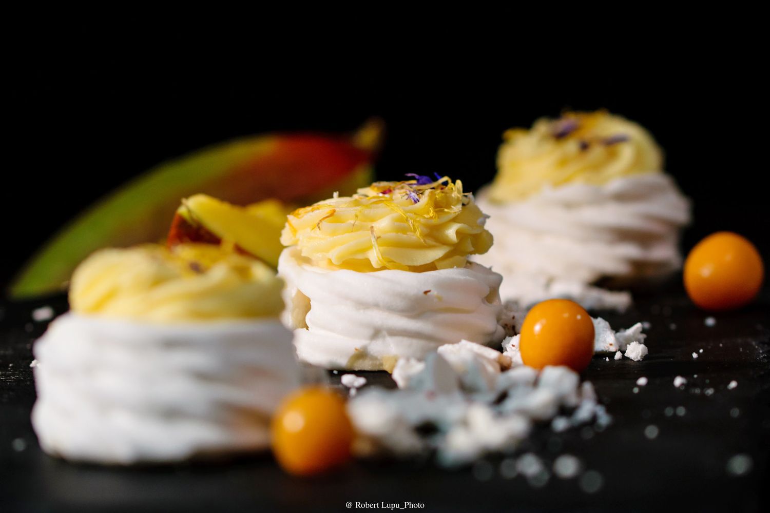 Delicate meringue nests filled with lemon cream and topped with kumquats on a dark surface.