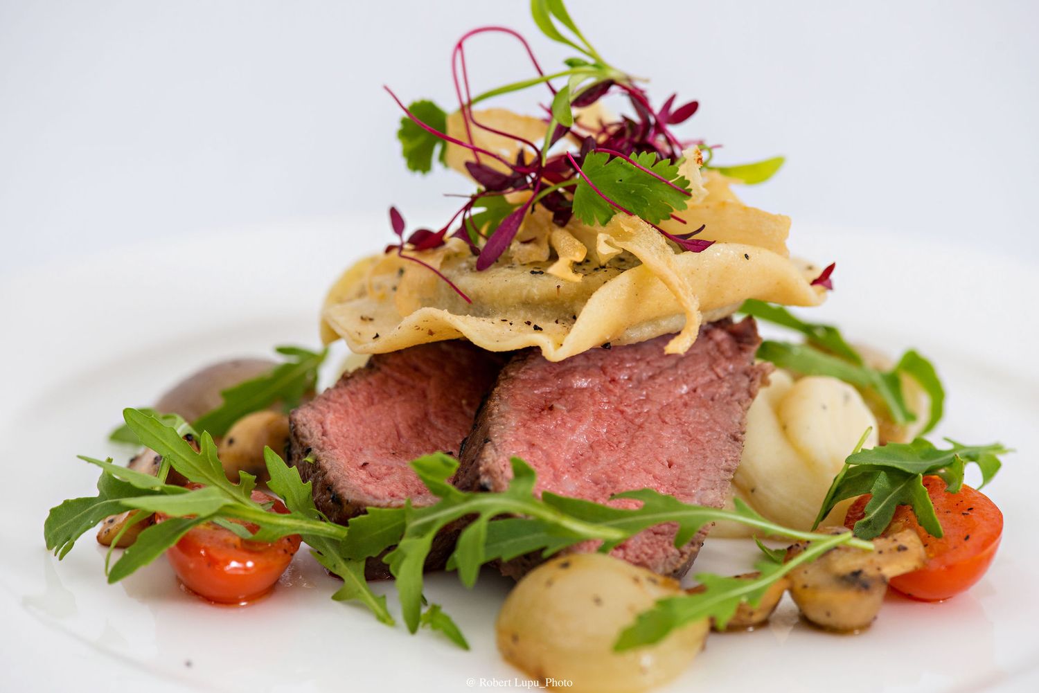 Elegant plated beef tenderloin with arugula, cherry tomatoes, mushrooms, and pasta crisps on a white dish.