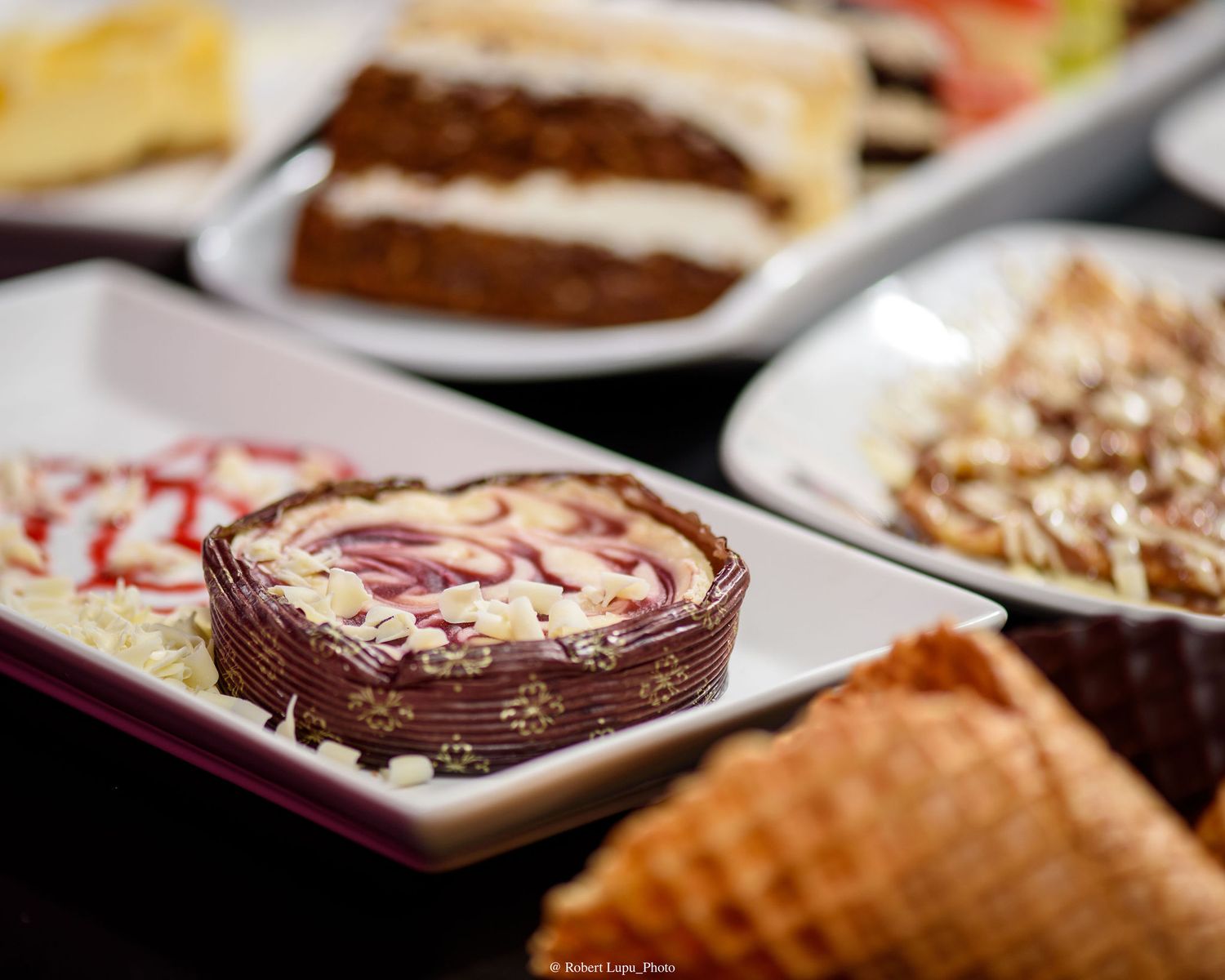 Assorted desserts on white plates including a chocolate-wrapped cheesecake slice and layered cake at a buffet spread.