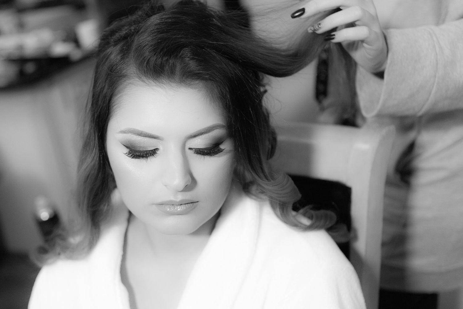 Black and white photo of a bride getting her hair styled while wearing a white robe, showcasing elegant makeup.
