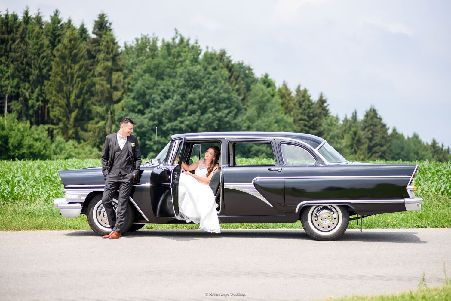 Bride and groom posing beside a classic black vintage car on a sunny day with green trees in the background.